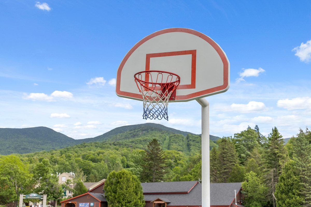 Summer at Smugglers' Notch Resort: a basketball hoop in front of green mountains with cut ski trails, and the resort welcome sign